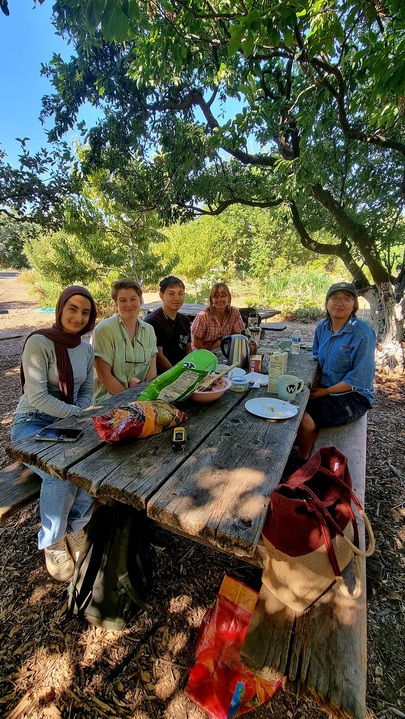 5 fresh focus team members sitting at a wooden picnic table in the ecological garden. There is food on the table left over from their meal, and they are smiling at the camera. 