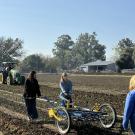 Fresh Focus students walk behind a Treffler handheld weeding device, weeding a bed of spinach. There is a tractor in the background. , 